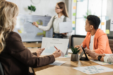 Focus on hands holding financial report. Female corporate director with laptop in hands having business meeting with international colleagues. Various diagrams, charts and technology at office.
