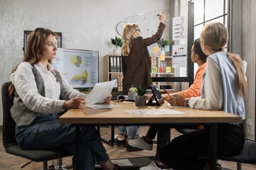 Caucasian mature woman writing financial report on glass board for multiracial female colleagues at modern office. Business women partners sitting at desk. Business conference concept.