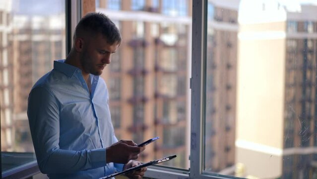 Focused Caucasian young man with document folder surfing Internet on smartphone looking out the window at residential multistorey building. Concentrated new flat owner on the left planning repair