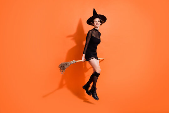 Full length photo of charming young lady sit broomstick soar wear stylish black halloween witch clothes isolated on orange color background