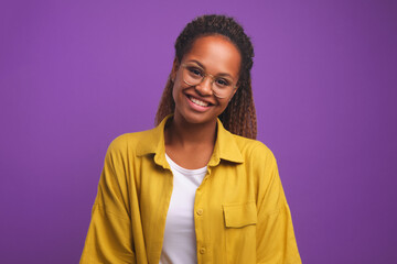 Young cheerful beautiful African American woman student wearing glasses smiling sincerely tilt head to side and look at you dressed in casual style stands on purple plain background