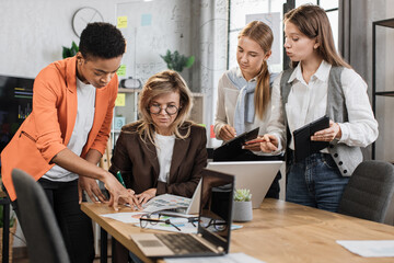 Teamwork, brainstorming in office. Diverse multiracial business people working together with laptop and tablet pc while their senior woman leader sign in report papers.