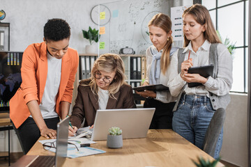 Teamwork, brainstorming in office. Diverse multiracial business people working together with laptop and tablet pc while their senior woman leader sign in report papers.