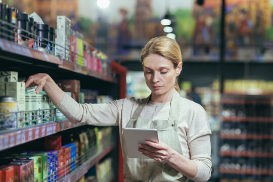A beautiful young worker in an apron is taking inventory in a supermarket, store. He stands with a notebook in his hands and takes a product from the shelf.