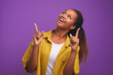 Young attractive ethnic African American woman looks up with delight and points with two fingers at something dressed in yellow shirt stands on purple plain background. Promotional offer