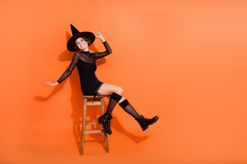Fototapeta na wymiar Full size photo of pretty young woman sit chair dangle legs wear stylish black halloween witch outfit isolated on orange color background