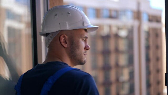 Close-up pensive Caucasian man in hard hat looking out the window at multistorey building in urban city. Side view of thoughtful builder in uniform standing indoors looking away thinking