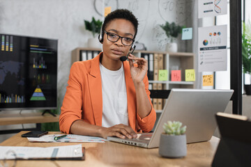 Portrait of attractive african business woman in headset sitting at office desk and looking at camera. Business lady having video conference showing thumb up. Business meeting online.
