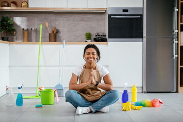 Glad young black lady sit on floor with mop, cleaning supplies meditating in white kitchen interior