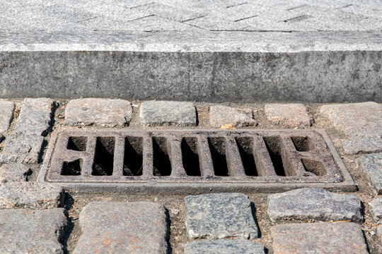 used iron rectangular manhole drainage system on the old cobbled road nearby to the granite curb and stone tile pedestrian pavement close-up on sunny dry weather, nobody.
