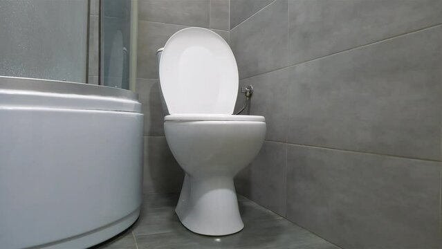 Toilet room interior with white toilet bowl with closed lid, shower cabin and grey wall tiles