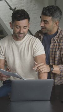 Gay Hispanic Couple Using Laptop And Looking At Paperwork In Kitchen