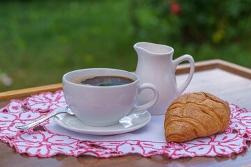 A cup of black coffee,croissant and milk on a table on natural background in garden.