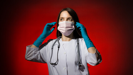 young woman doctor in a white coat and medical gloves puts on a medical mask on a red background
