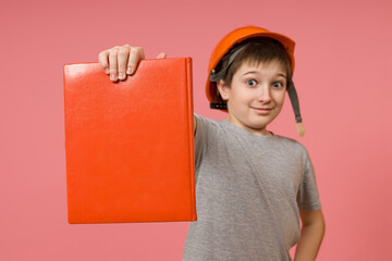 a boy in a construction helmet and a gray t-shirt shows a book to the camera and stands on a pink...