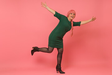 funny young woman in a pink wig and green dress stands on one leg with her hands up on a colored background