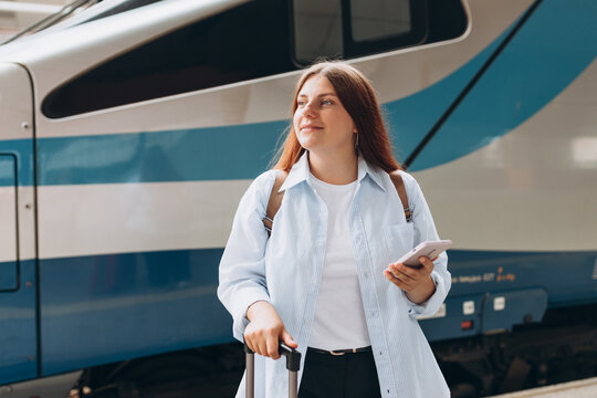 Young redhhead woman waiting train with backpack and using smart phone. Railroad transport concept, Traveler.Woman with suitcase walking at railroad station platform. Travel to vacation by train