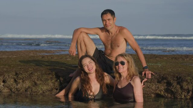 Handsome man sitting on a reef stone and two women bathing in ocean water posing and smiling for the picture on beach at sunset. Happy time for a family spending time together. Taking photo for memory