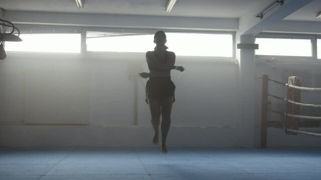Sportswoman skipping rope, intensive cardio training in gym, front view wide shot. Young professional fighter silhouette exercising, jumping