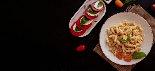  Italian pasta fusilli and caprese salad with tomato, mozzarella and basil with pesto on black rustic background. Extra wide banner. Copy space