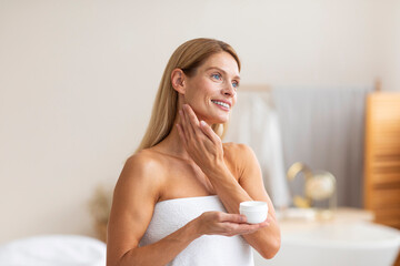 Daily skincare. Beautiful middle aged female applying cream and holding jar with moisturizer,...