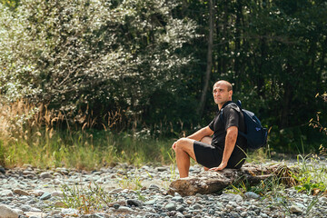 a male traveler sits on a log against the backdrop of a green forest, rests and enjoys the view of nature