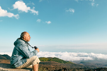 Naklejka na ściany i meble Mature bearded man looking through binoculars at panoramic view of colorful summits of active volcano Etna, Tallest volcano in Continental Europe, Sicily, Italy.