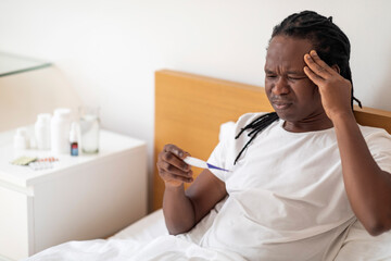 Sick Young African American Man Holding Thermometer While Sitting In Bed