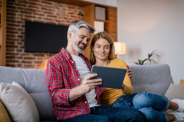 Cheerful mature european couple have video call remotely, chatting by tablet sit on sofa