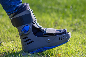 Boy with broken foot and orthopedic shoe or walker after bone fracture rests in public park on...