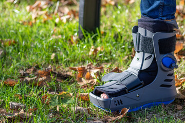 Boy with broken foot and orthopedic shoe or walker after bone fracture rests in public park on...