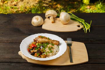 Rice with stewed vegetables and baked chicken leg on a handmade wooden board next to mushrooms and green onions.