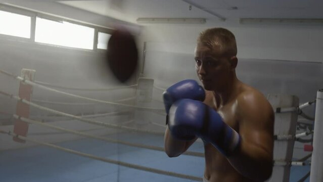 Boxer training with punchbag in ring, handheld tracking shot. Young fighter exercising with professional equipment in gym, hitting boxing pear