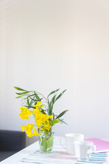 Shrub branch with Tecoma stans flowers in a glass beaker on a white table in the interior. A pink spring notepad, two coffee mugs and a geometric patterned napkin.