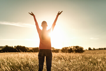 Young woman with arms outstretched up the sunset sky standing in a field feeling free and happy  - obrazy, fototapety, plakaty