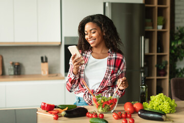 Smiling pretty young african american lady preparing salad, reading message on smartphone in kitchen interior
