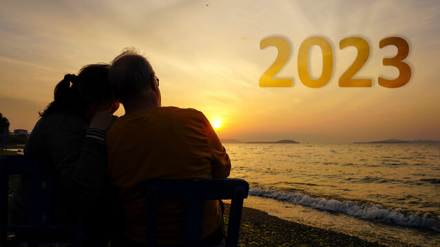 Happy New Year 2023 is coming concept. Old couple watching beautiful yellow sunset or sunrise on beach. High resolution photo can be used as large display, print, website banner, social media post.