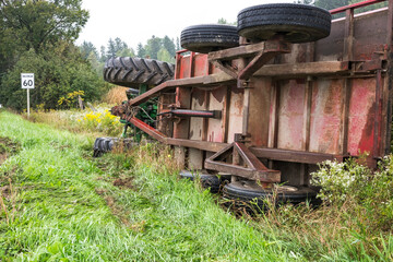 An overturned farm tractor and cart sit in a ditch at the side of a road.