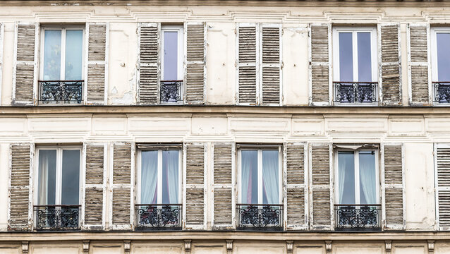 Wood shutters with peeling paint flank windows with black wrought iron railings on a building in Paris France.