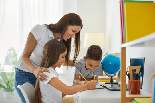 Happy mother watches her preschool son and daughter write or draw together at home. Happy young Caucasian woman and her children are studying and doing creative work. Family concept.
