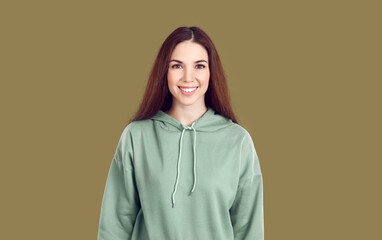 Portrait of smiling teen girl in hoodie isolated on green khaki studio background feel positive and joyful. Profile picture of happy young Caucasian woman in casual wear look at camera.