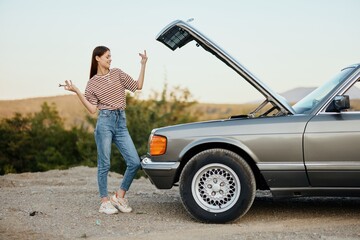 A woman stands outside a broken-down, dangerous old car with a wrench on the road traveling alone