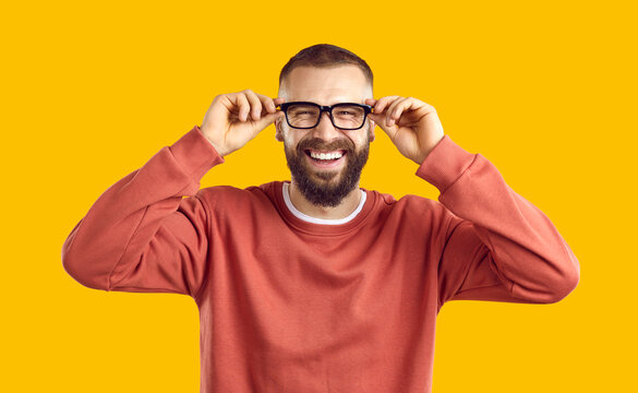 Cheerful, Smiley, Funny Man Puts On His Eyeglasses. Studio Shot Of Happy, Laughing Handsome Bearded Young Guy Wearing Glasses And Orange Sweatshirt Standing Isolated On Yellow Background