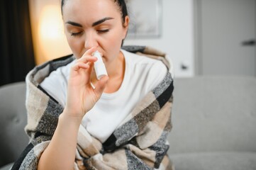 Nasal spray in hand of sick young woman sitting on couch. Allergic rhinitis symptoms and treatment