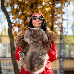 A girl in sunglasses, a headscarf and a red jacket holds a British cat in her arms on an autumn background. Walk with a cat. Love to the animals. Autumn mood. Blurred background.