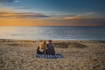 Teenage boy and younger teenage girl with long red hair sit on blanket on sandy beach watching sunset; sea and colorful sky in background