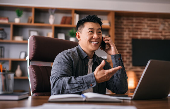 Smiling handsome adult chinese male at table with laptop speaks by phone with client in office interior