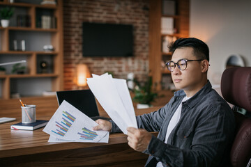 Serious concentrated adult japanese man investor in glasses works with papers, charts and graphs in dark office