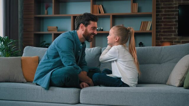Playful caucasian young family daddy and small daughter sitting comfortable on couch loving cute girl touching father nose funny joke playing game with parent having fun spending free time together