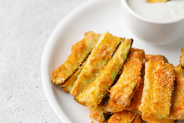 Zucchini sticks baked with cheese, herbs, breadcrumbs and cream yogurt sauce. Roasted Zucchini snack on white plate.